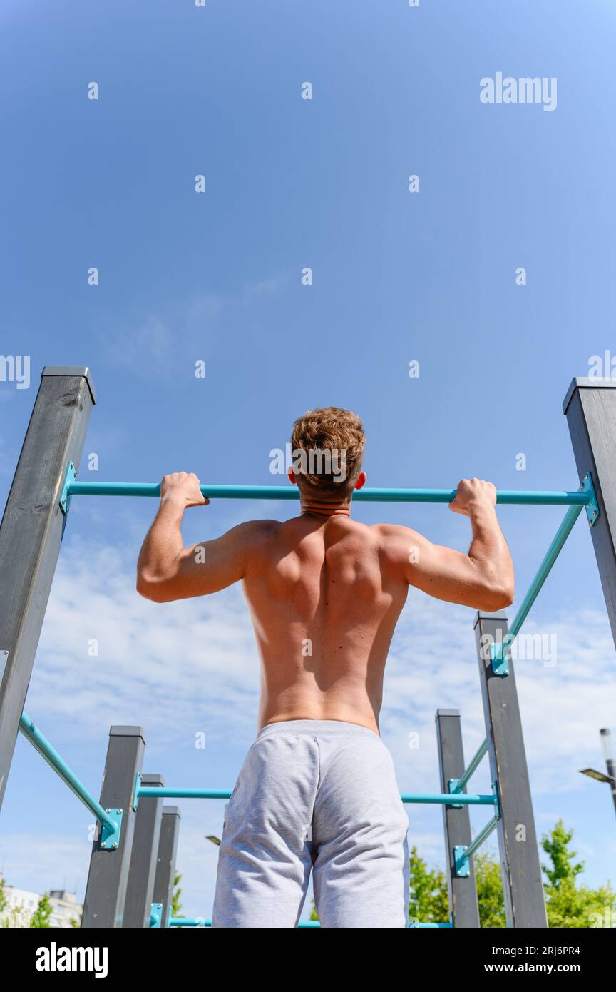Un giovane con un torso nudo si tira su un bar orizzontale in un campo sportivo di strada per un allenamento. Vista dal retro. Cielo blu sullo sfondo Foto Stock