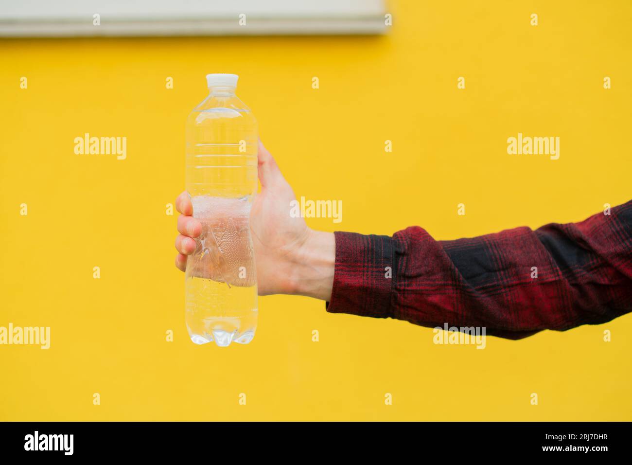 Primo piano uomo irriconoscibile con una camicia rossa tiene tra le mani una bottiglia d'acqua trasparente davanti a uno sfondo giallo della parete Foto Stock