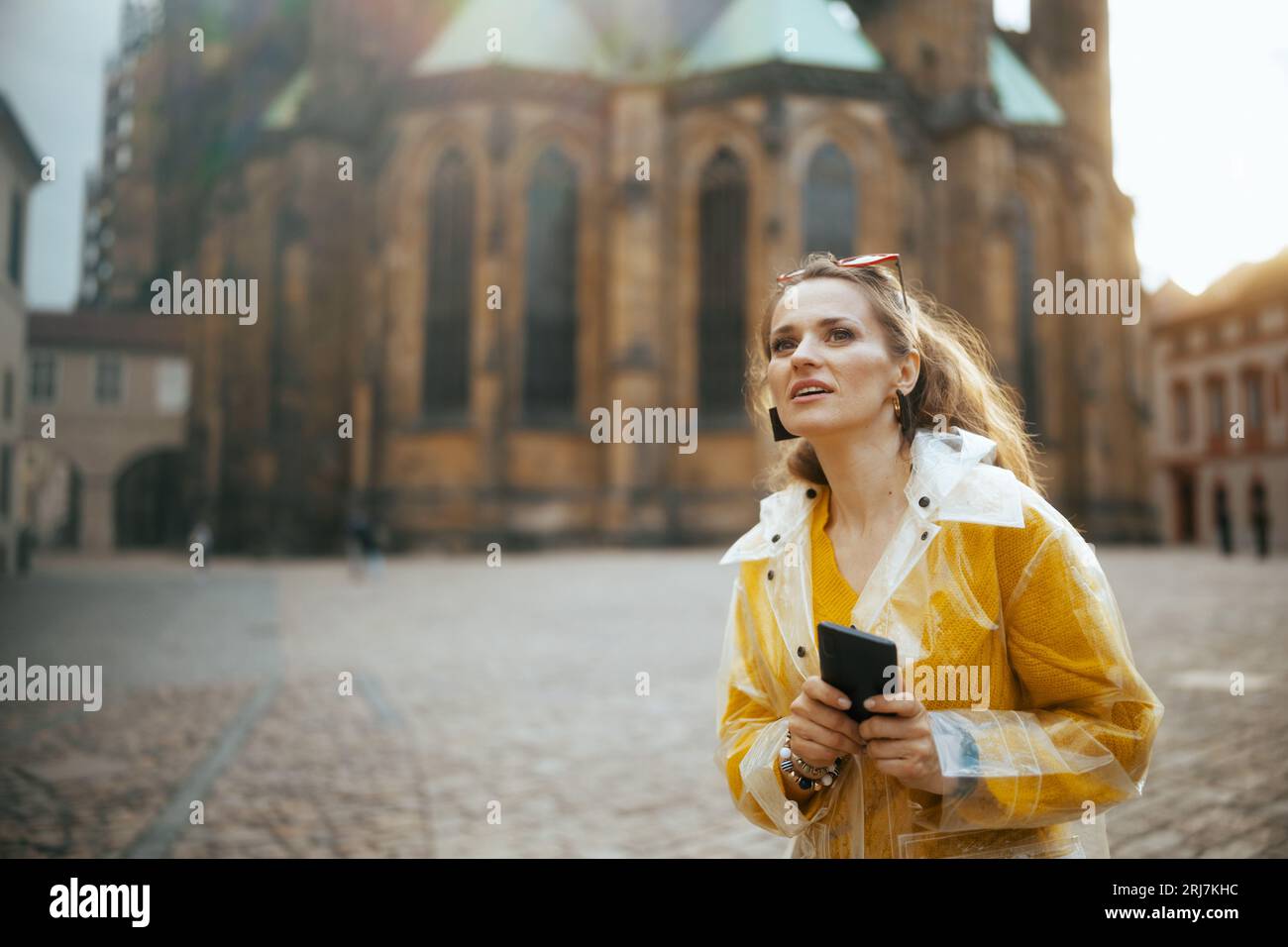 Una donna viaggiatrice moderna sorpresa con camicetta gialla e impermeabile a Praga Repubblica Ceca che ha fatto un tour a piedi e utilizza lo smartphone. Foto Stock