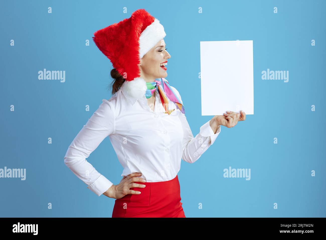 Elegante hostess sorridente su sfondo blu in uniforme con cappello di Babbo Natale che mostra un foglio di carta a4. Foto Stock