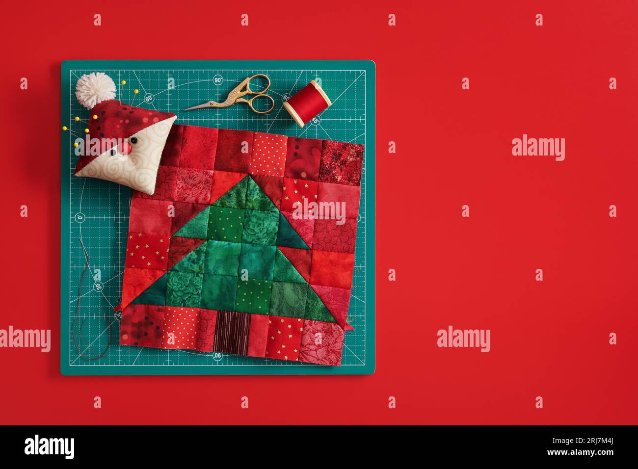 Blocco patchwork albero di Natale, cuscinetto come Babbo Natale, forbici, bobina di filo su tappetino artigianale, sfondo rosso Foto Stock