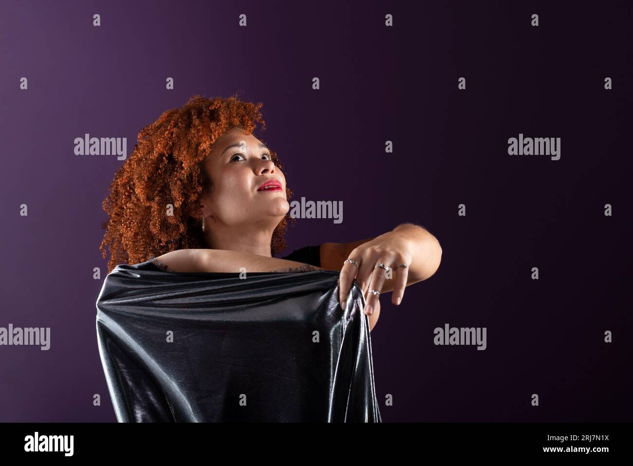 Bella e attraente donna con capelli rossi ricci, con un panno argentato. Indossa abiti neri. Isolato su sfondo lilla colorato. Foto Stock