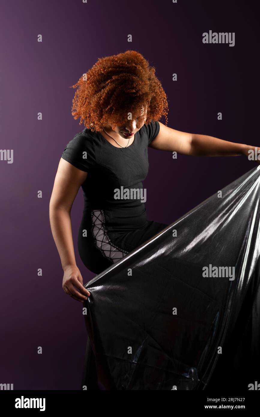 Bella e attraente donna con capelli rossi ricci, con un panno argentato. Indossa abiti neri. Isolato su sfondo lilla colorato. Foto Stock