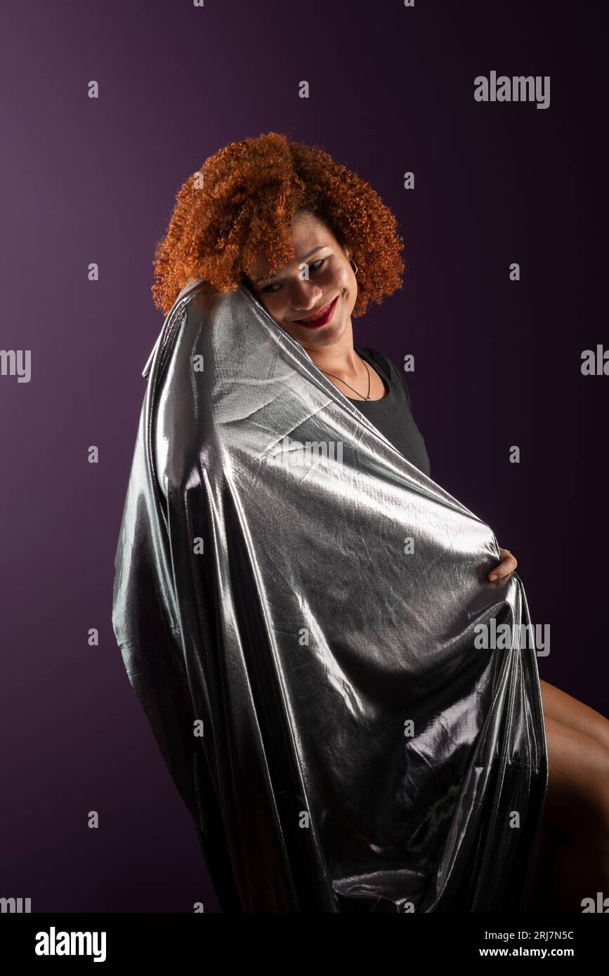 Bella e attraente donna con capelli rossi ricci, con un panno argentato. Indossa abiti neri. Isolato su sfondo lilla colorato. Foto Stock