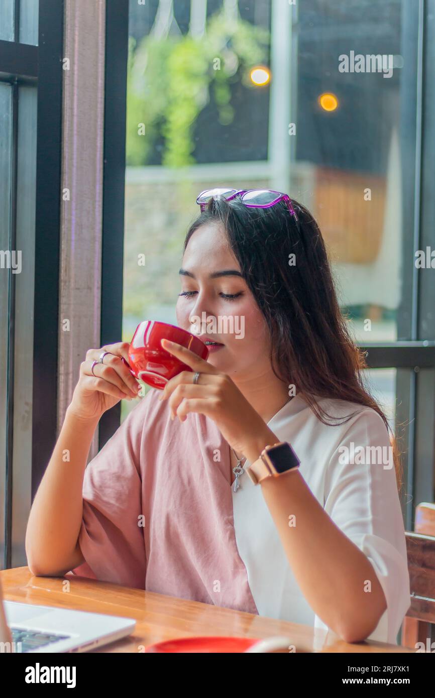 giovane donna asiatica che tiene una tazza di caffè e si ferma. Bella giovane donna d'affari asiatica al bar, che usa il computer portatile e beve caffè, sorridendo Foto Stock