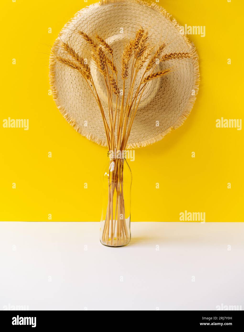 Foto di natura naturale del grano in bottiglia di vetro davanti al cappello del contadino e alla parete gialla. Foto Stock