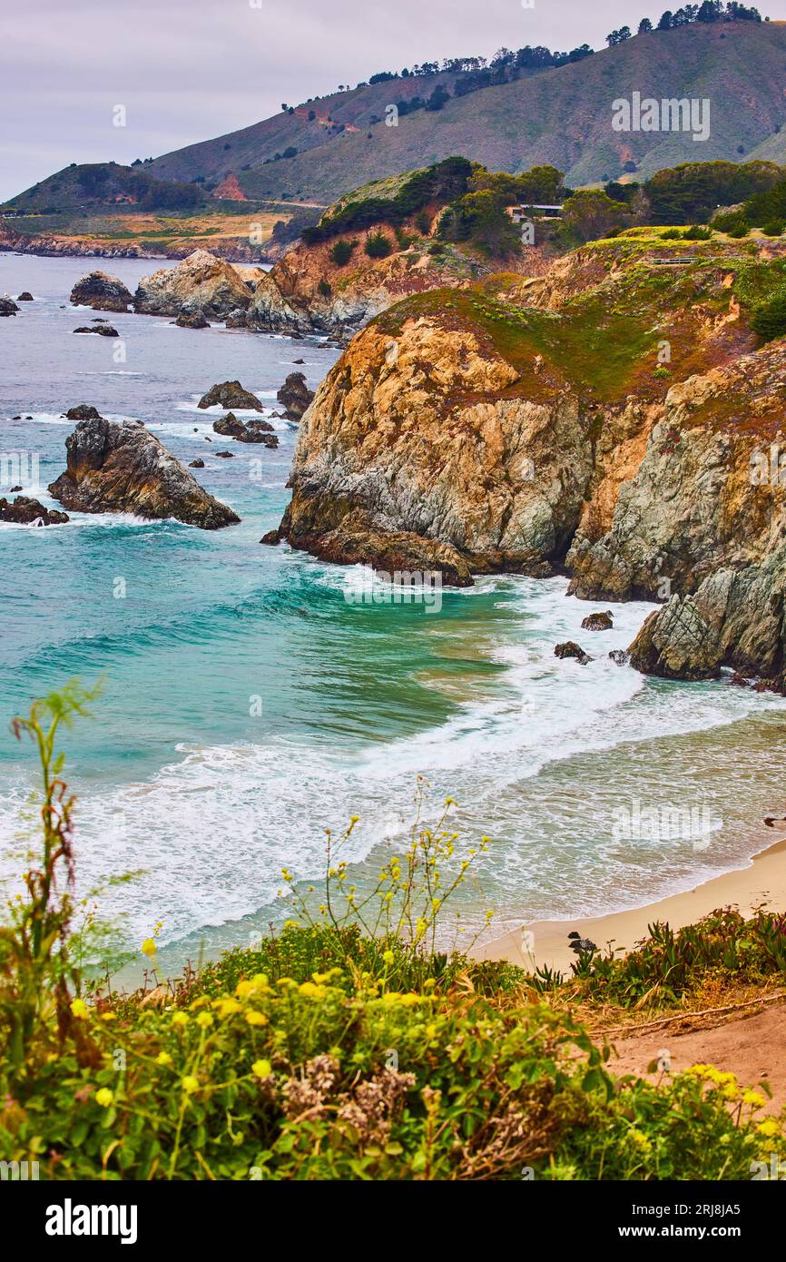 Le splendide onde di color tè si infrangono in una baia sabbiosa con pareti di scogliera e lontane colline montuose Foto Stock