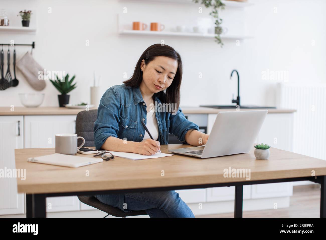 Adulti coreani concentrati che scrivono informazioni su carta mentre scorrono le pagine Web sul laptop nella cucina moderna. Donna consapevole in abbigliamento casual che raccoglie fatti Foto Stock
