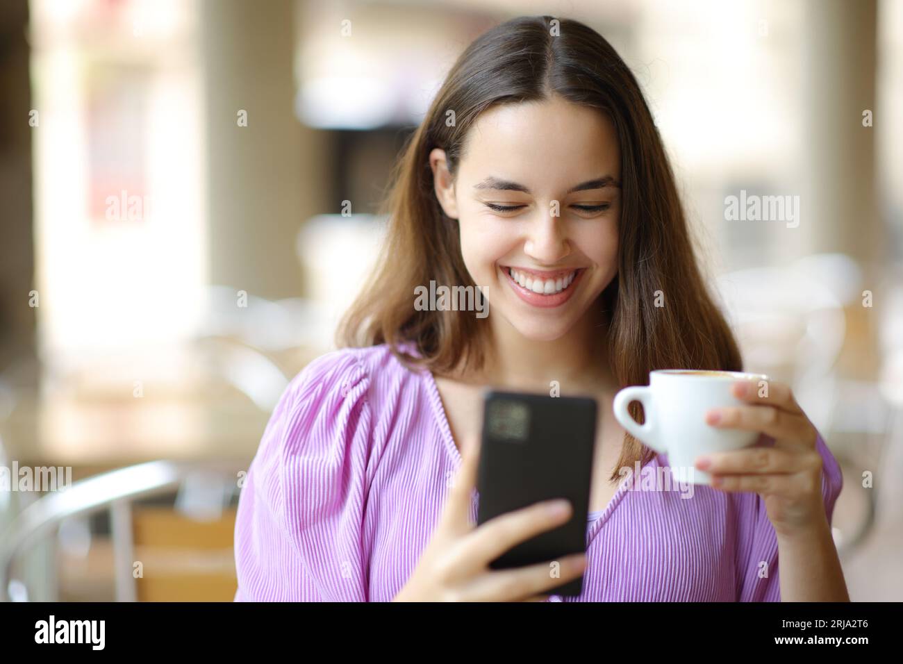 Donna felice che usa il telefono bevendo caffè seduto in una terrazza del ristorante Foto Stock