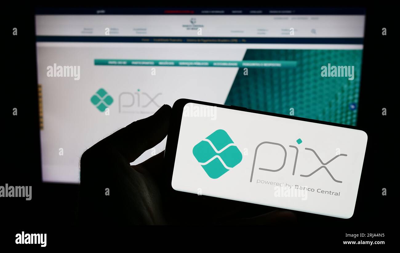 Persona che possiede uno smartphone con il logo della piattaforma brasiliana di pagamento istantaneo Pix sullo schermo di fronte al sito Web. Concentrarsi sul display del telefono. Foto Stock