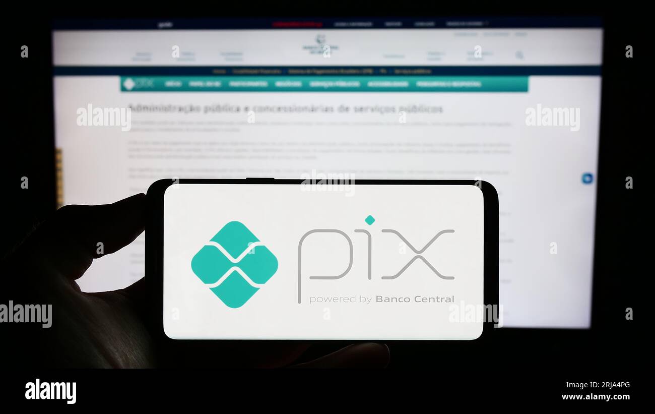 Persona che possiede il cellulare con il logo della piattaforma brasiliana di pagamento istantaneo Pix sullo schermo di fronte alla pagina Web aziendale. Concentrarsi sul display del telefono. Foto Stock