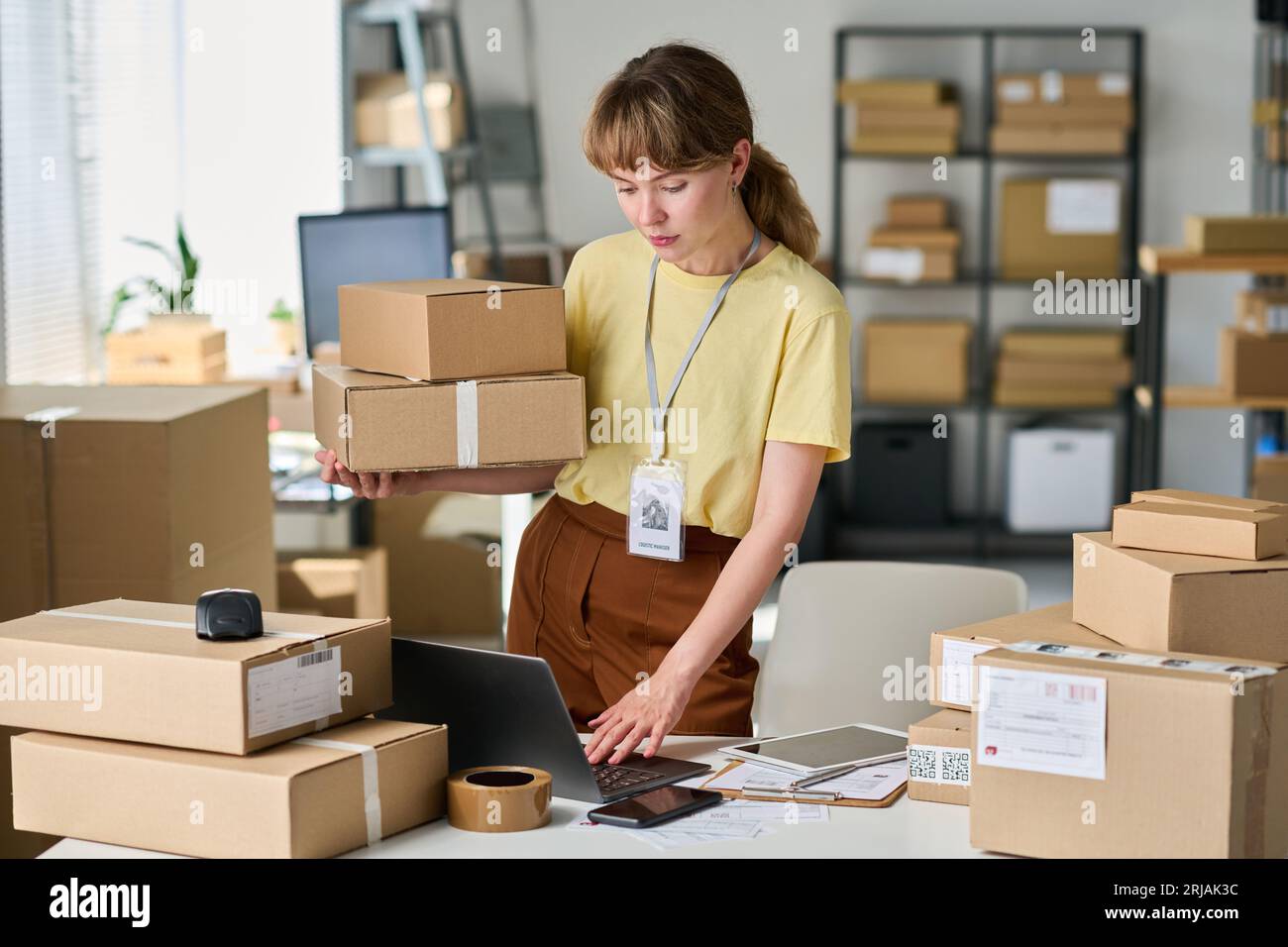 Giovane donna in abbigliamento casual che tiene una pila di scatole imballate e utilizza un computer portatile mentre controlla i dati personali dei clienti nel database Foto Stock