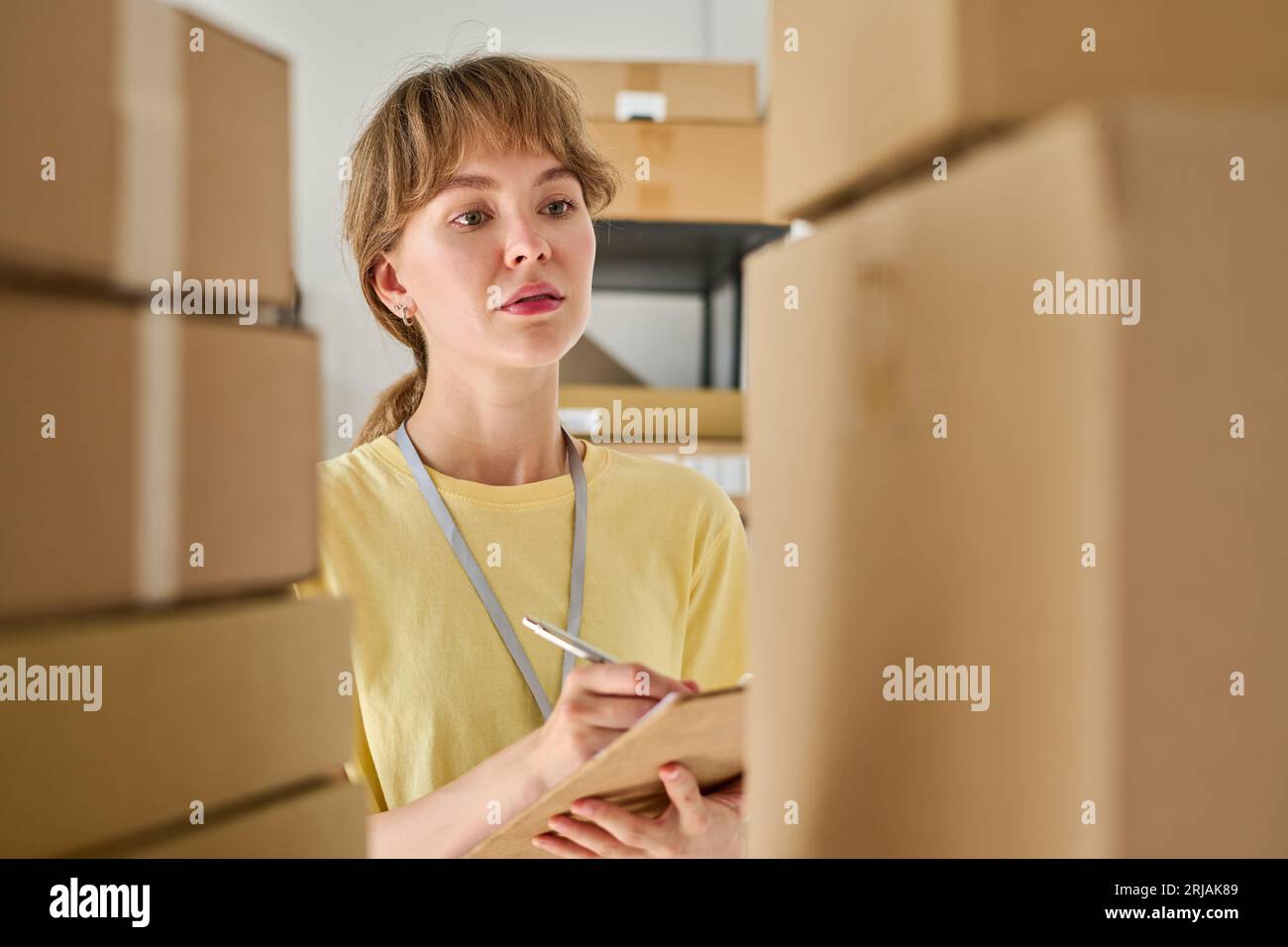 Giovane donna seria con t-shirt gialla che prende appunti nel documento mentre guarda una pila di scatole di cartone confezionate in piedi sullo scaffale Foto Stock