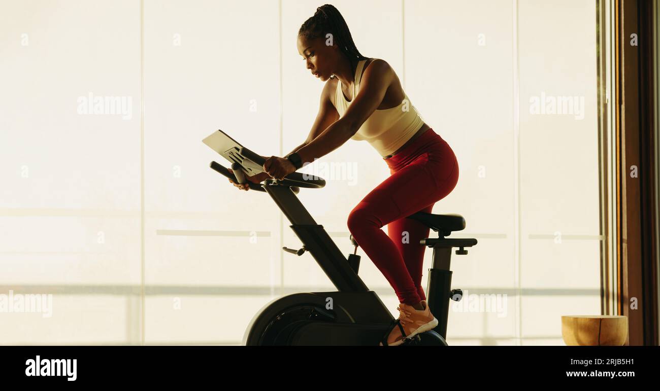 Donna africana in forma che lavora su una cyclette digitale, impegnandosi in cardio per mantenere la sua salute e il suo benessere. Donna che usa attrezzature intelligenti per l'esercizio fisico Foto Stock