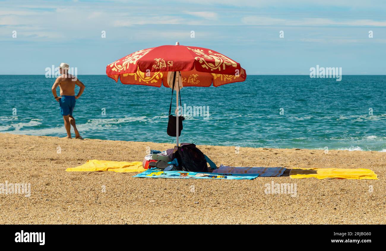 L'uomo si trova vicino al mare vicino a un ombrellone che ripara gli effetti personali, Nazare, Portogallo Foto Stock