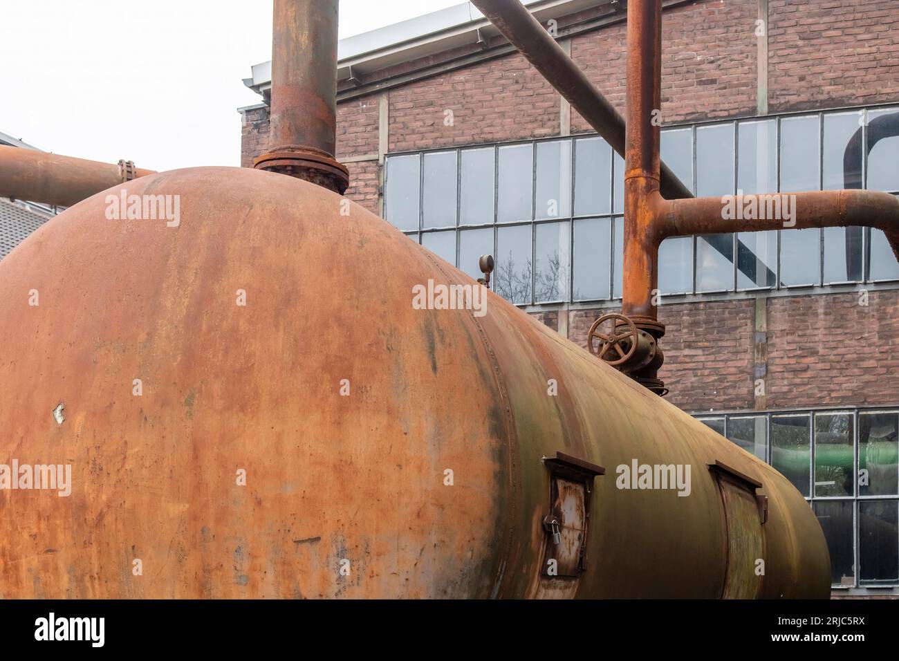 Primo piano ad angolo ridotto di serbatoio arrugginito e intemprato con sistema di tubazioni per liquidi di fronte all'edificio in un acciaio industriale abbandonato e fatiscente m Foto Stock