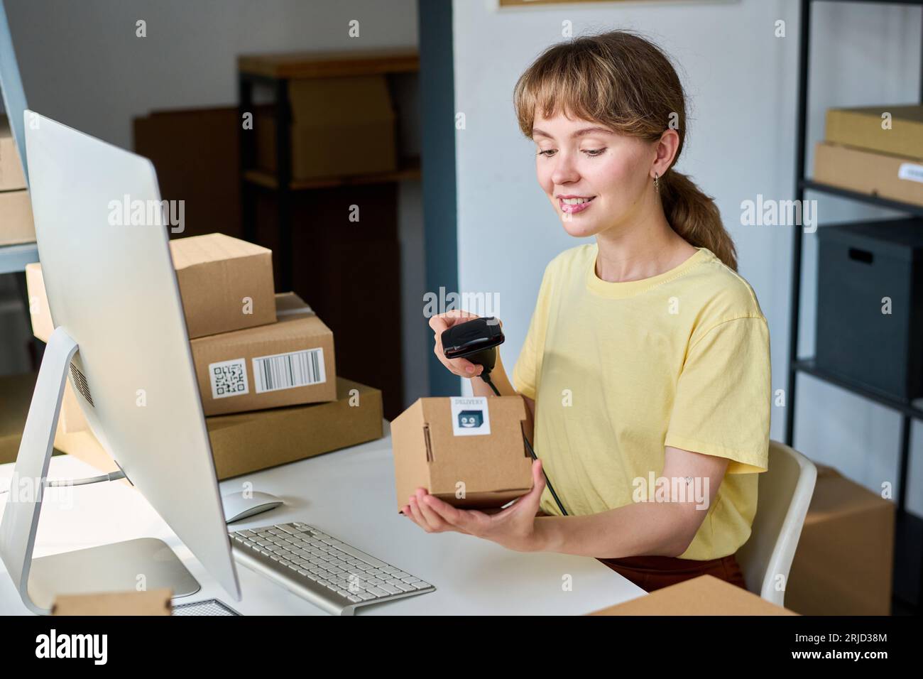 Giovane donna sorridente che scansiona la scatola con lo scanner mentre è seduta sul posto di lavoro davanti al computer e controlla il codice qr sulla confezione Foto Stock