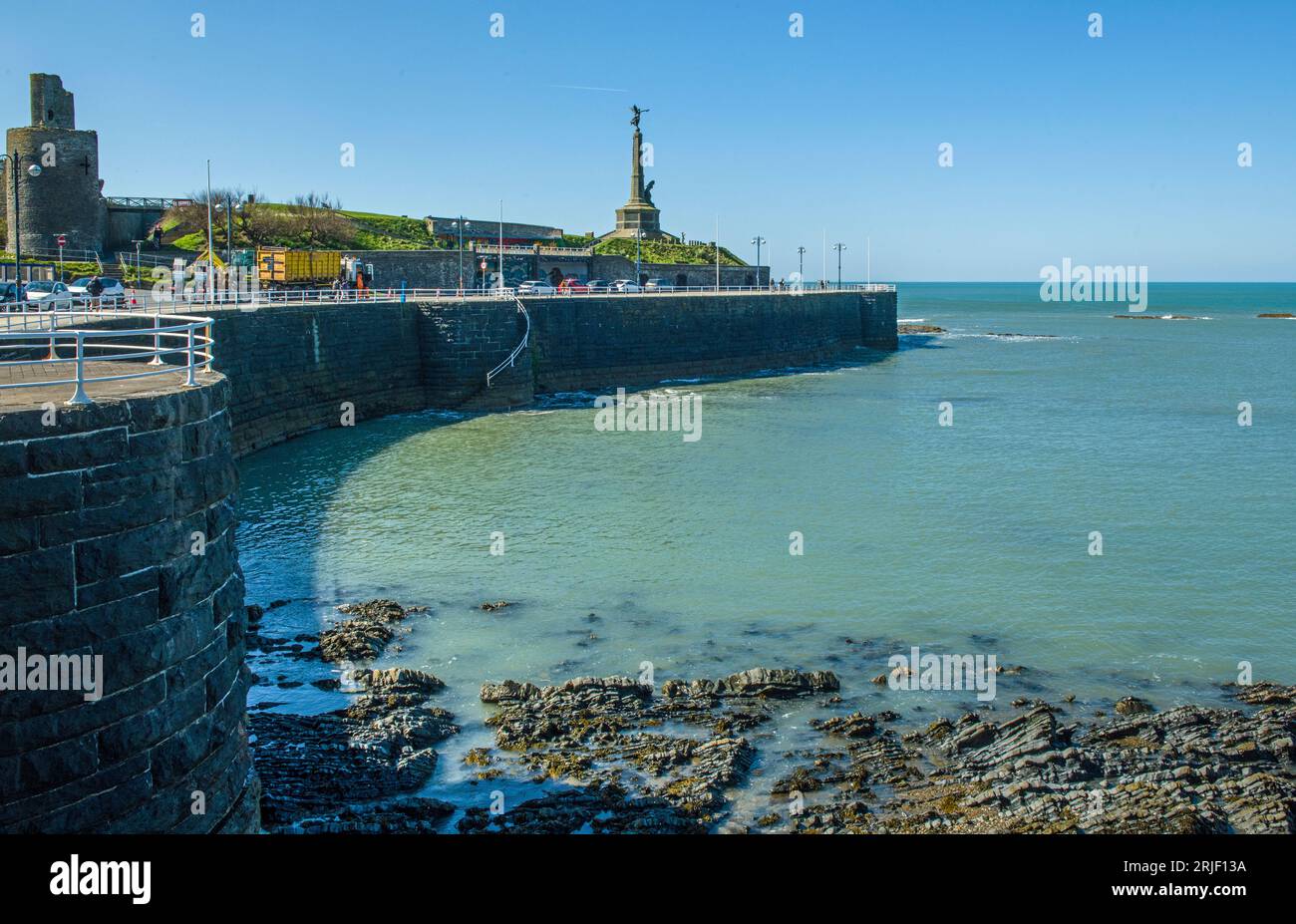 Il fronte ad Aberystwyth, soleggiato giorno di aprile, lungo la costa, con un muro di difesa del mare, un monumento, ringhiere e parte del vecchio castello Foto Stock