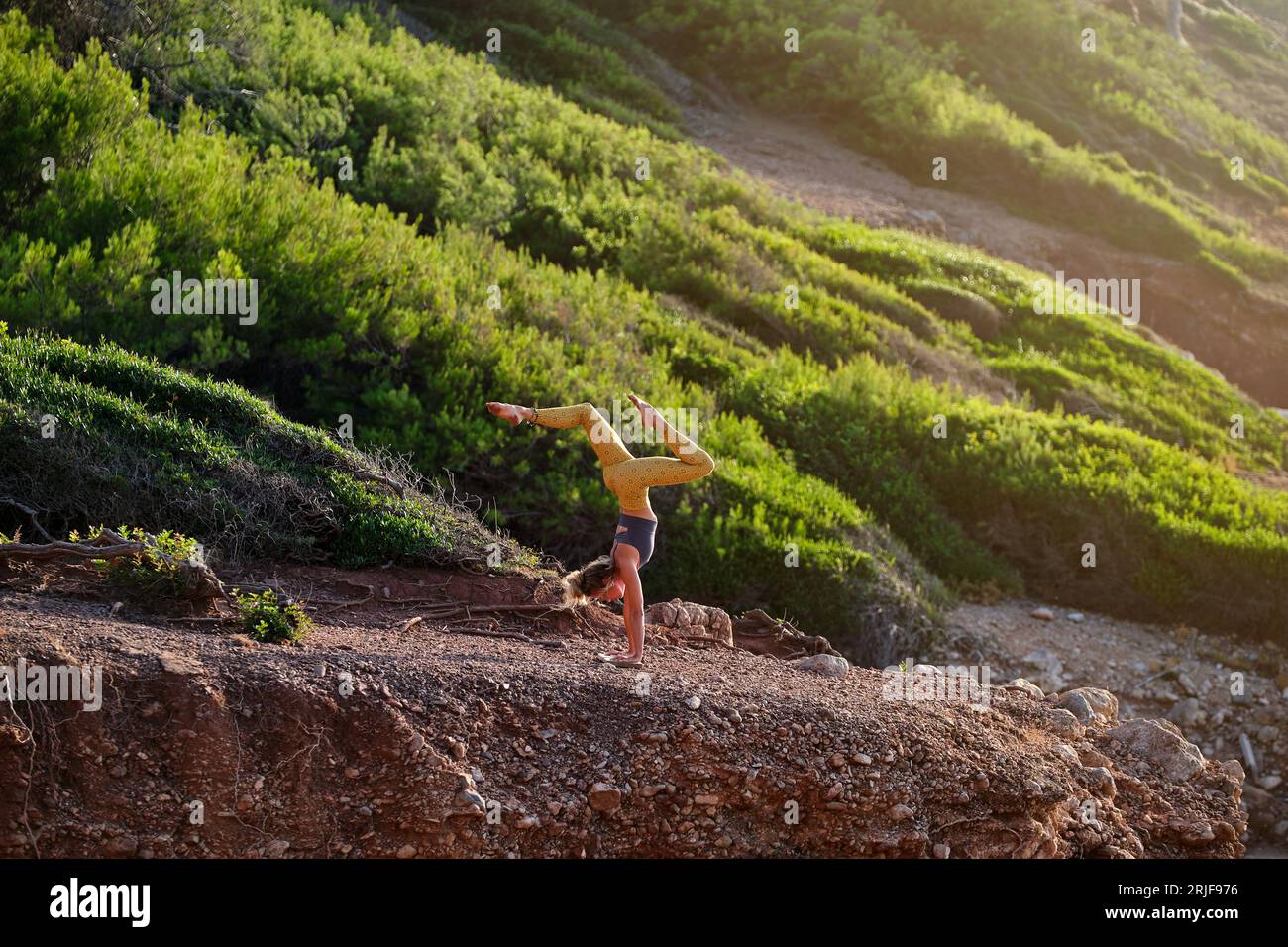 Vista laterale di una giovane atleta in abbigliamento sportivo che porta Adho Mukha Vrikshasana in posa con le gambe separate mentre fa yoga sulle piste in montagna Foto Stock