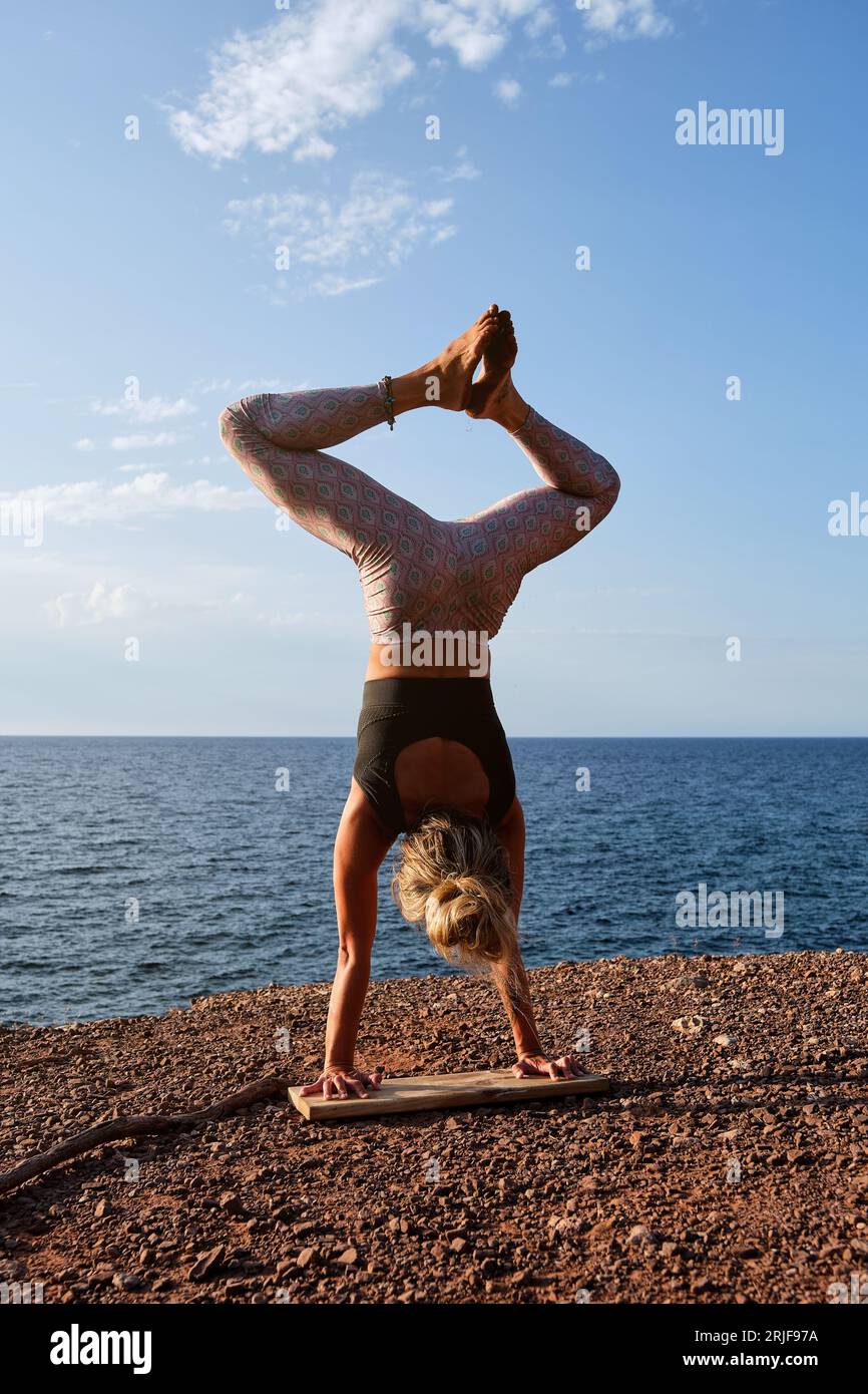 Vista posteriore di una atleta senza volto in abbigliamento sportivo in piedi su una tavola di legno in Adho Mukha Vrikshasana posa con gambe in Baddha Konasana mentre si pratica Foto Stock