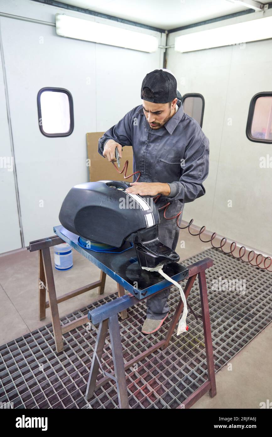 Un uomo serio che indossa indumenti da lavoro e tinteggiatura di berretti, serbatoio carburante per moto con pistola a spruzzo in garage Foto Stock