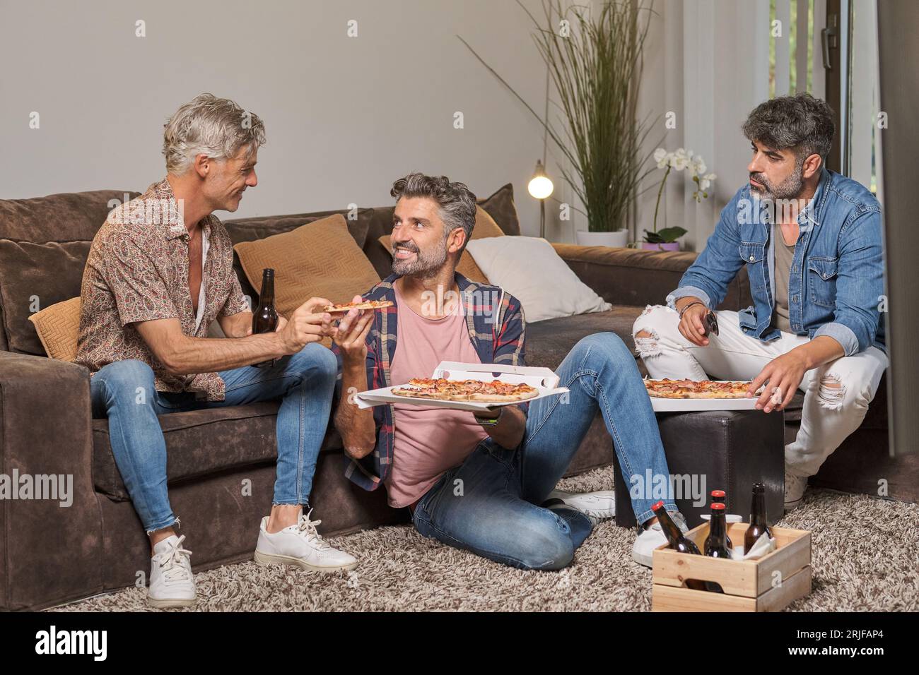 Uomini felici e maturi in abiti informali che mangiano una pizza gustosa mentre si divertono in salotto a casa Foto Stock