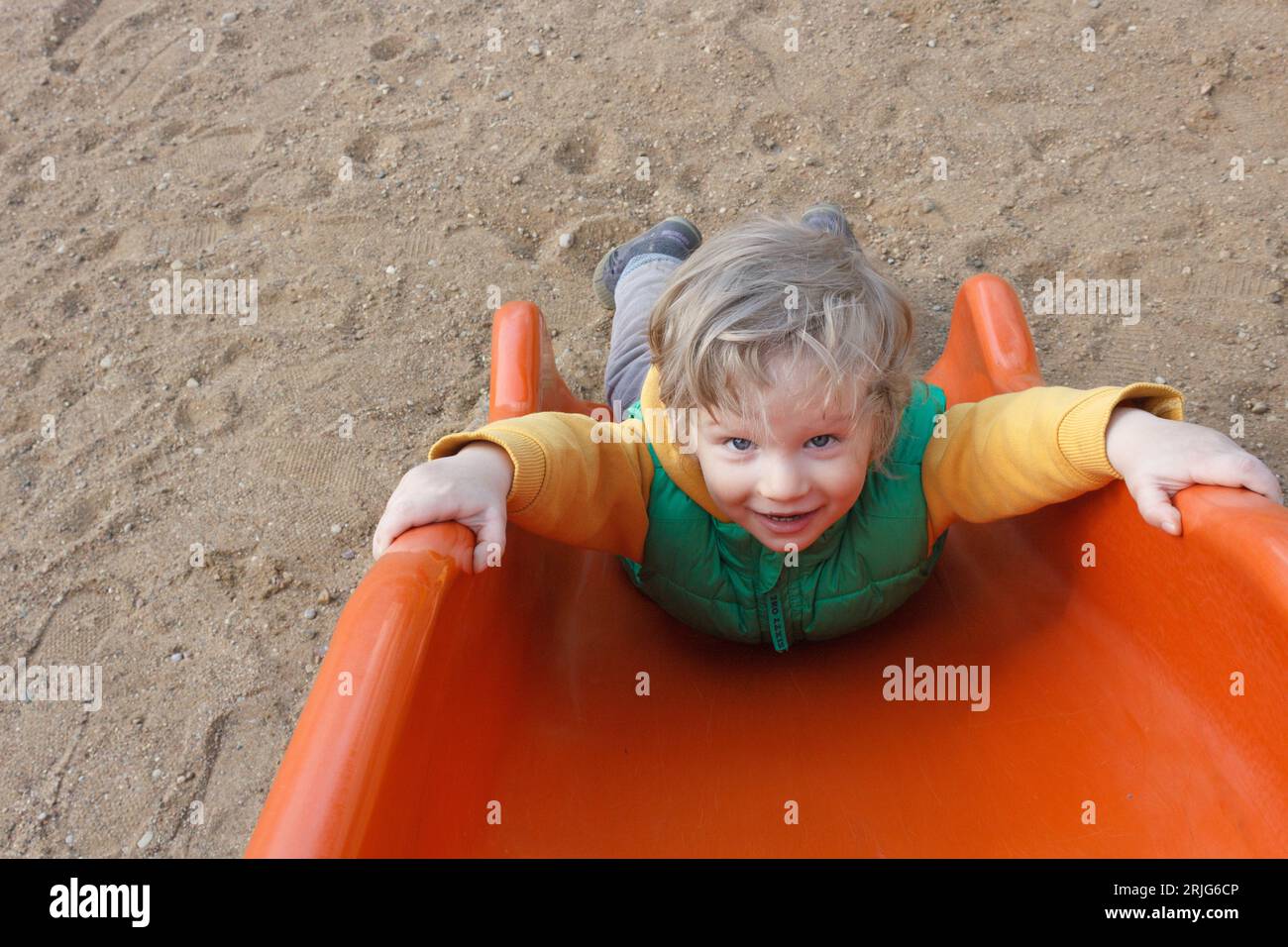 Un bambino felice gioca nel parco giochi - scivola lungo lo scivolo arancione. Foto Stock