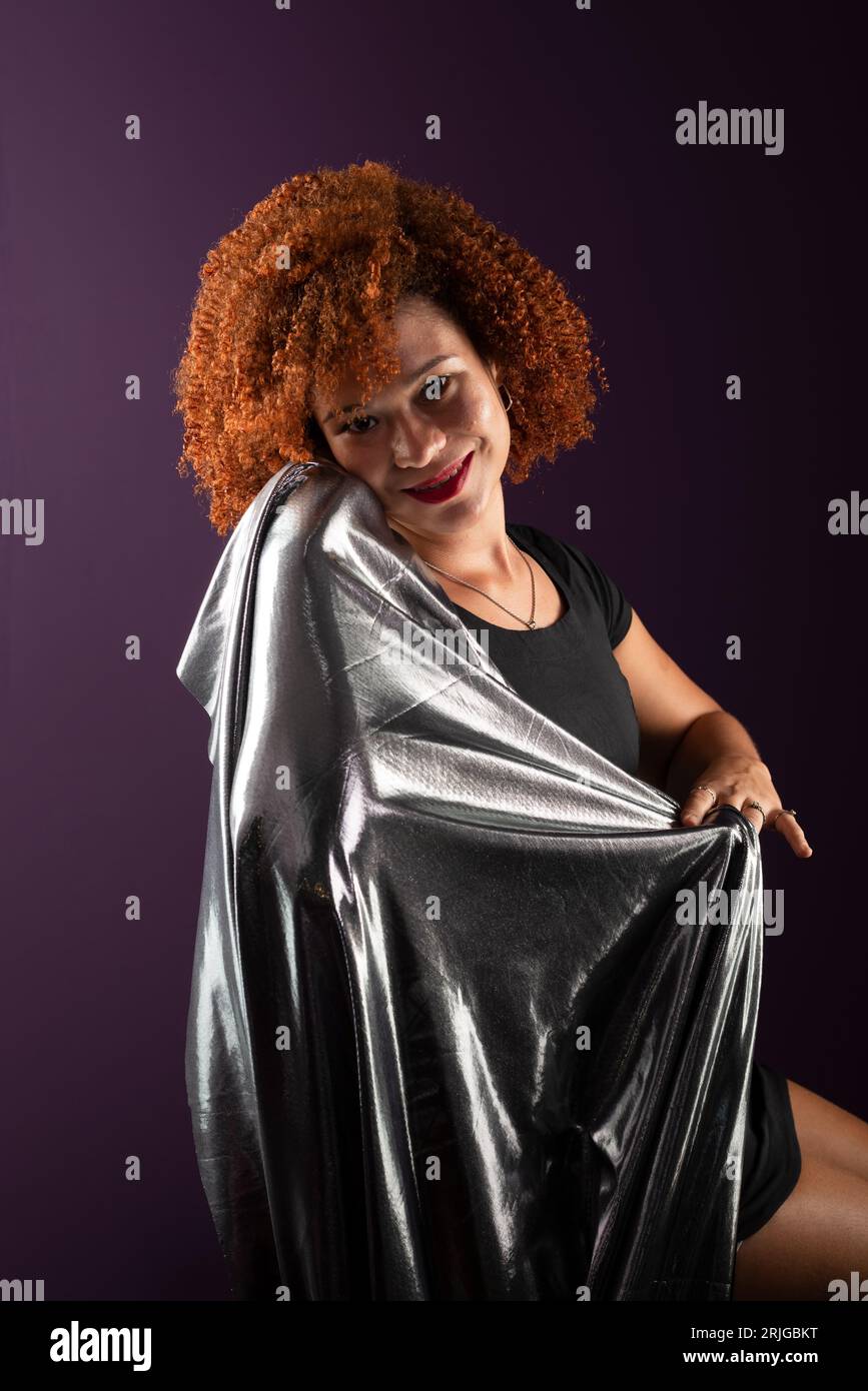 Bella e attraente donna con capelli rossi ricci, con un panno argentato. Indossa abiti neri. Isolato su sfondo lilla colorato. Foto Stock