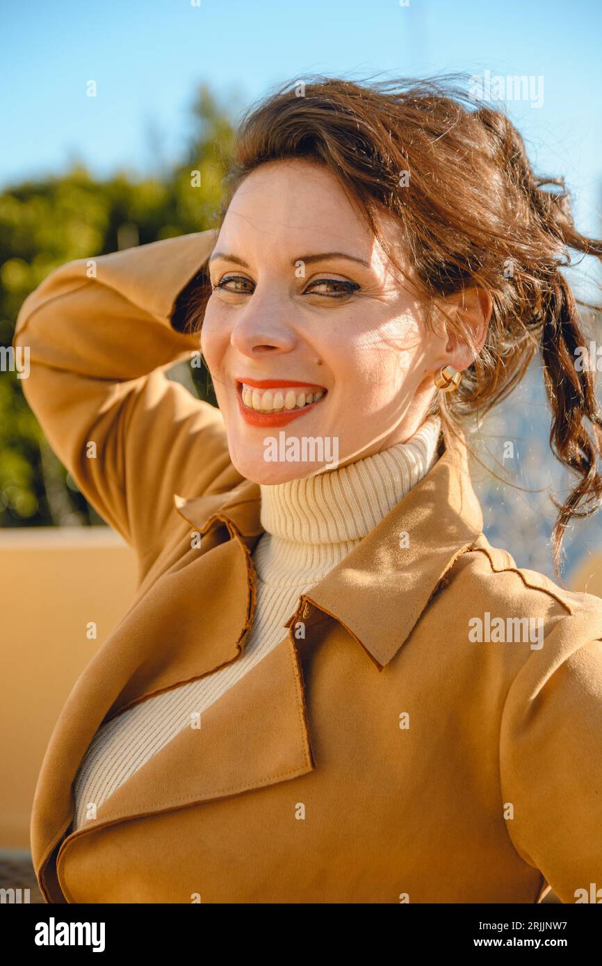 ritratto verticale di giovane ed elegante donna caucasica francese con labbra rosse e giacca marrone in piedi sulla terrazza al tramonto sorridendo felice e guardando ca Foto Stock