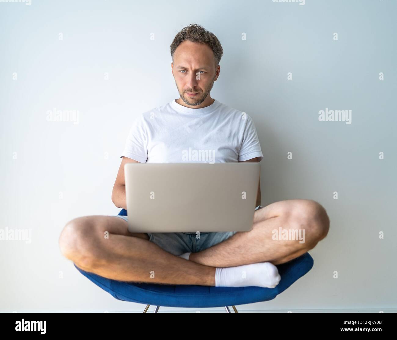 L'uomo lavora al computer portatile seduto su una sedia e a gambe incrociate. Freelance o lavoratore remoto che utilizza il computer a casa Foto Stock