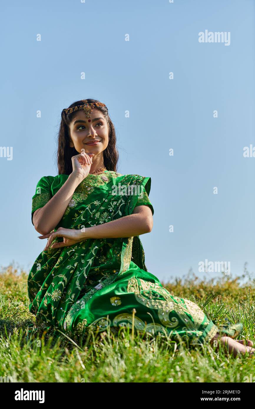 donna indiana positiva in sari tradizionali in posa su una collina erbosa con il cielo sullo sfondo Foto Stock