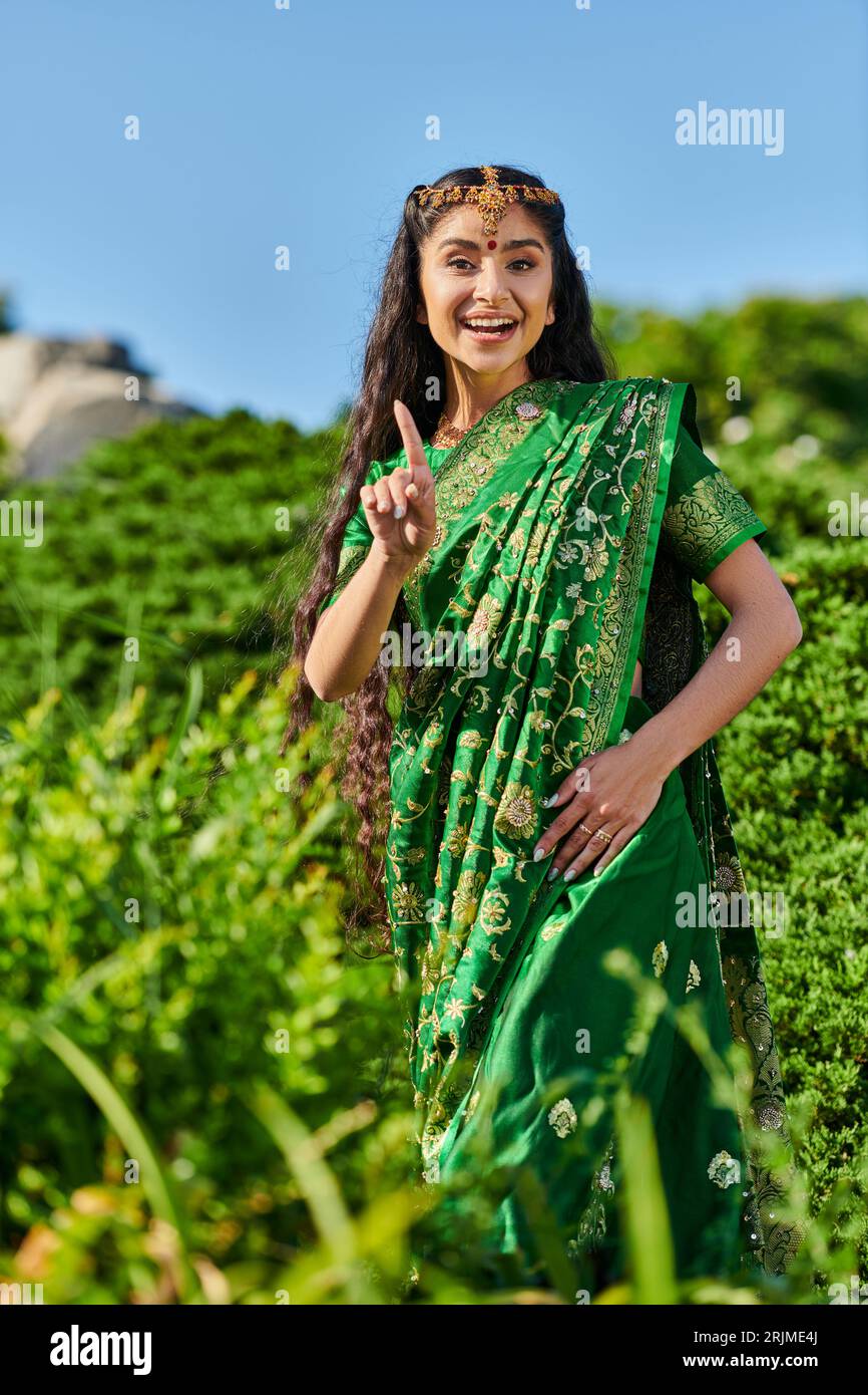 allegra giovane donna indiana in sari che punta con il dito mentre si trova vicino alle piante nel parco Foto Stock