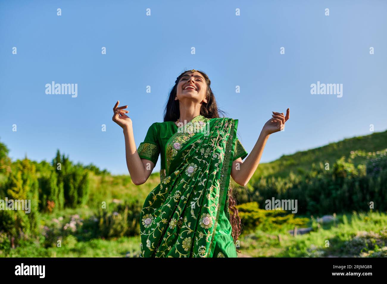 spensierata donna indiana in sari e matha patti mentre si trova in piedi con paesaggio estivo sullo sfondo Foto Stock