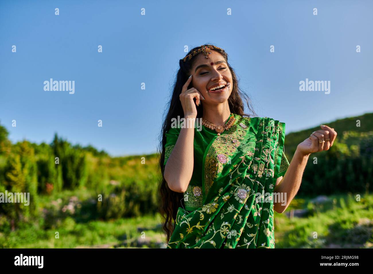 allegra giovane donna indiana che tocca il volto mentre posa in sari con il paesaggio sullo sfondo Foto Stock