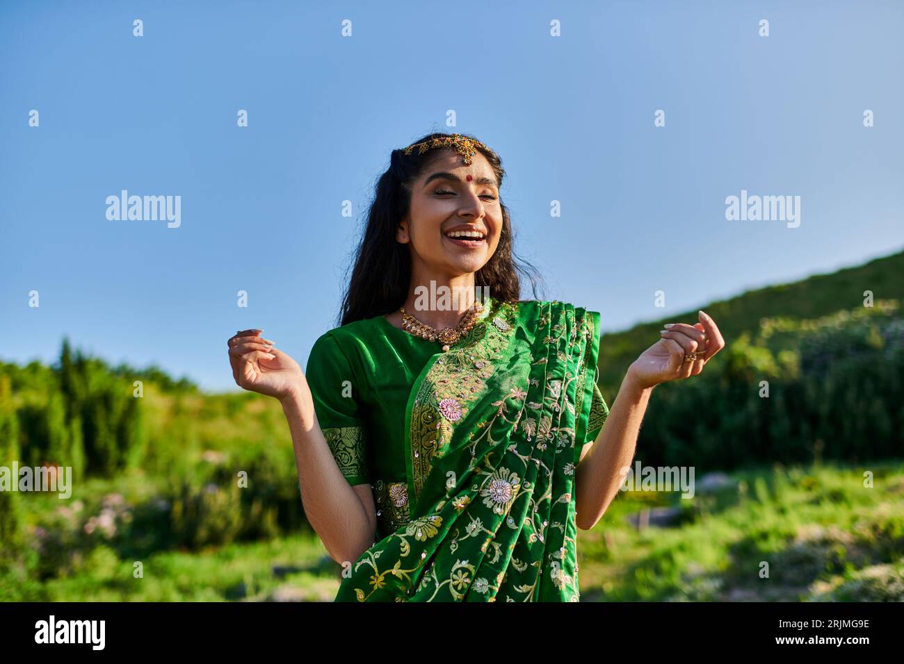 felice giovane donna indiana in elegante sari in posa e in piedi con paesaggio sullo sfondo Foto Stock
