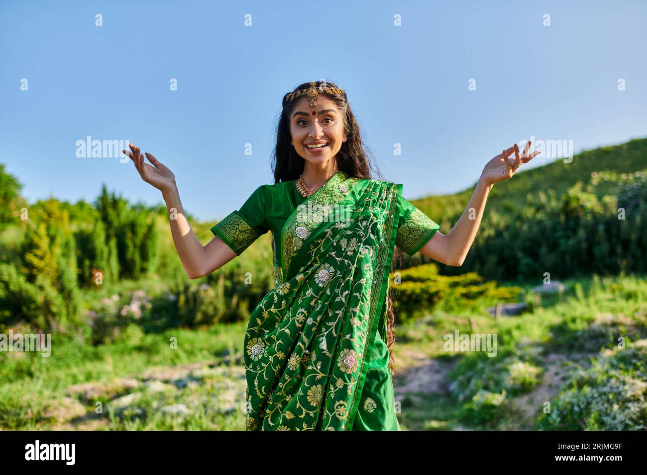 allegra giovane donna indiana in sari verdi che posa e guarda la macchina fotografica sul campo estivo Foto Stock