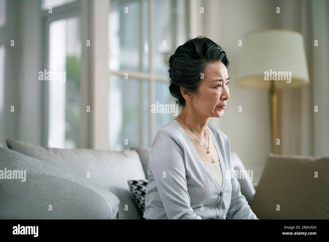 vista laterale di una donna asiatica anziana e triste seduta da sola sul divano in salotto a casa Foto Stock