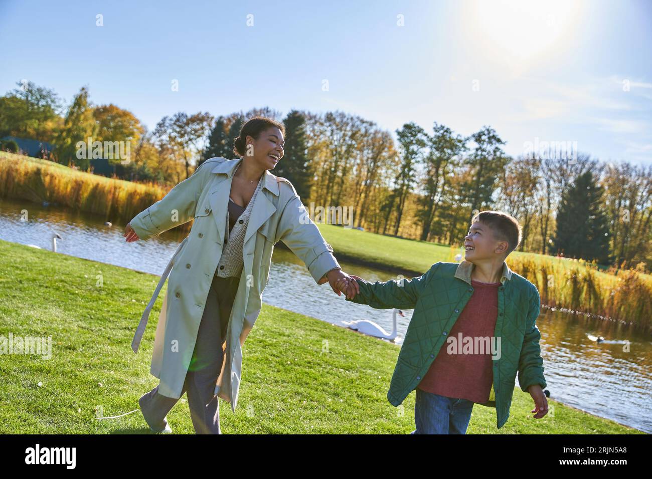 felicità, madre afroamericana che segue il figlio, cammina vicino allo stagno, genitorialità moderna, moda autunnale Foto Stock