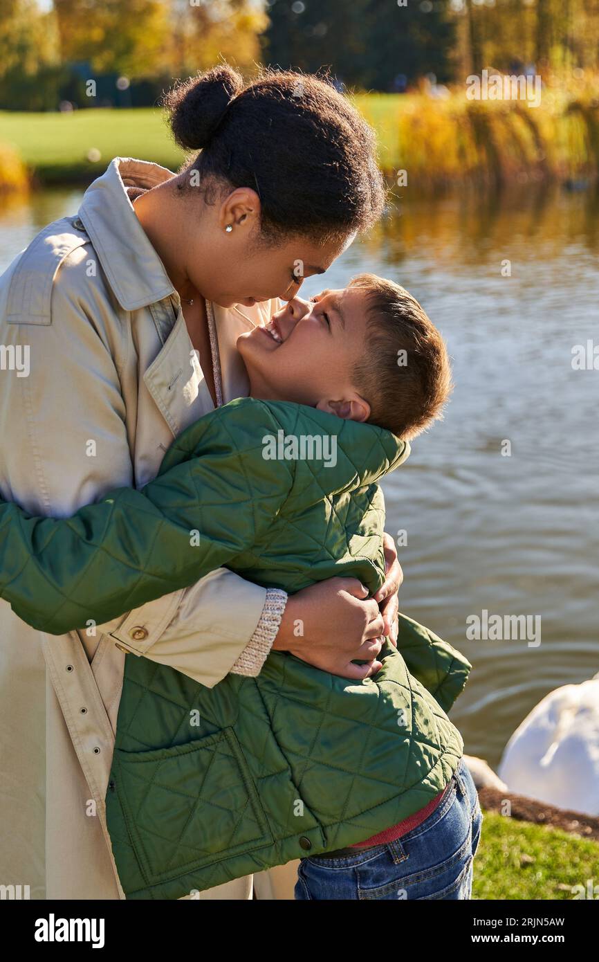 amore materno, ragazzo allegro che abbraccia la madre vicino al lago, famiglia afroamericana, autunno, autunno Foto Stock