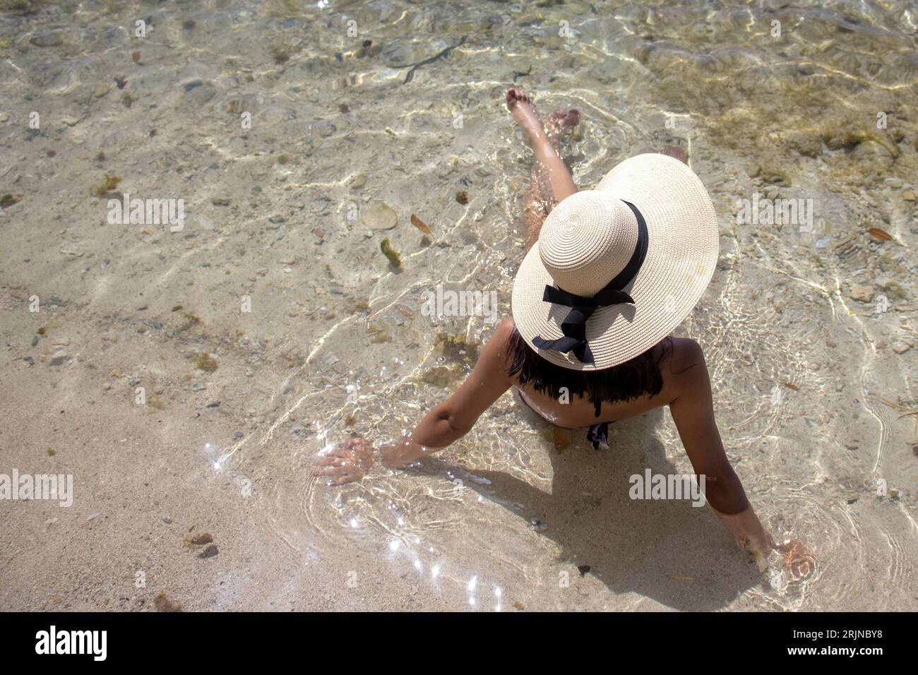 Una giovane donna con un elegante cappello e un bikini in due pezzi seduto in acque turchesi poco profonde Foto Stock