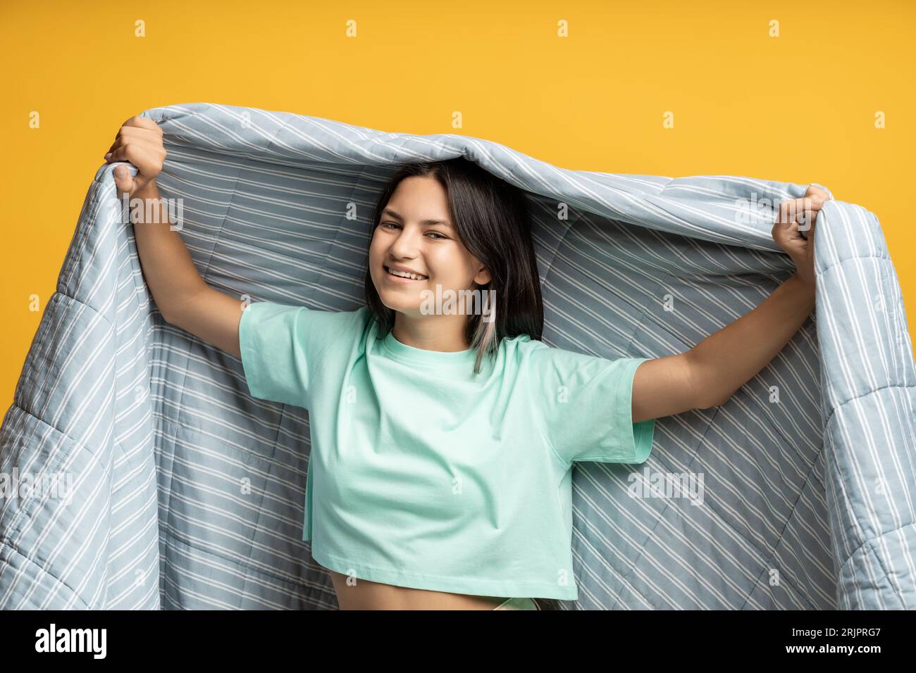 Adolescente sorridente in coperta isolata sul giallo. Happy Girl gode di un'intimità confortevole con un morbido piumone Foto Stock