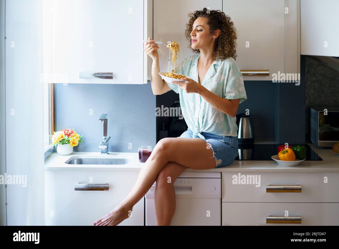 Donna sorridente che beve la pasta e si siede sul bancone della cucina Foto Stock