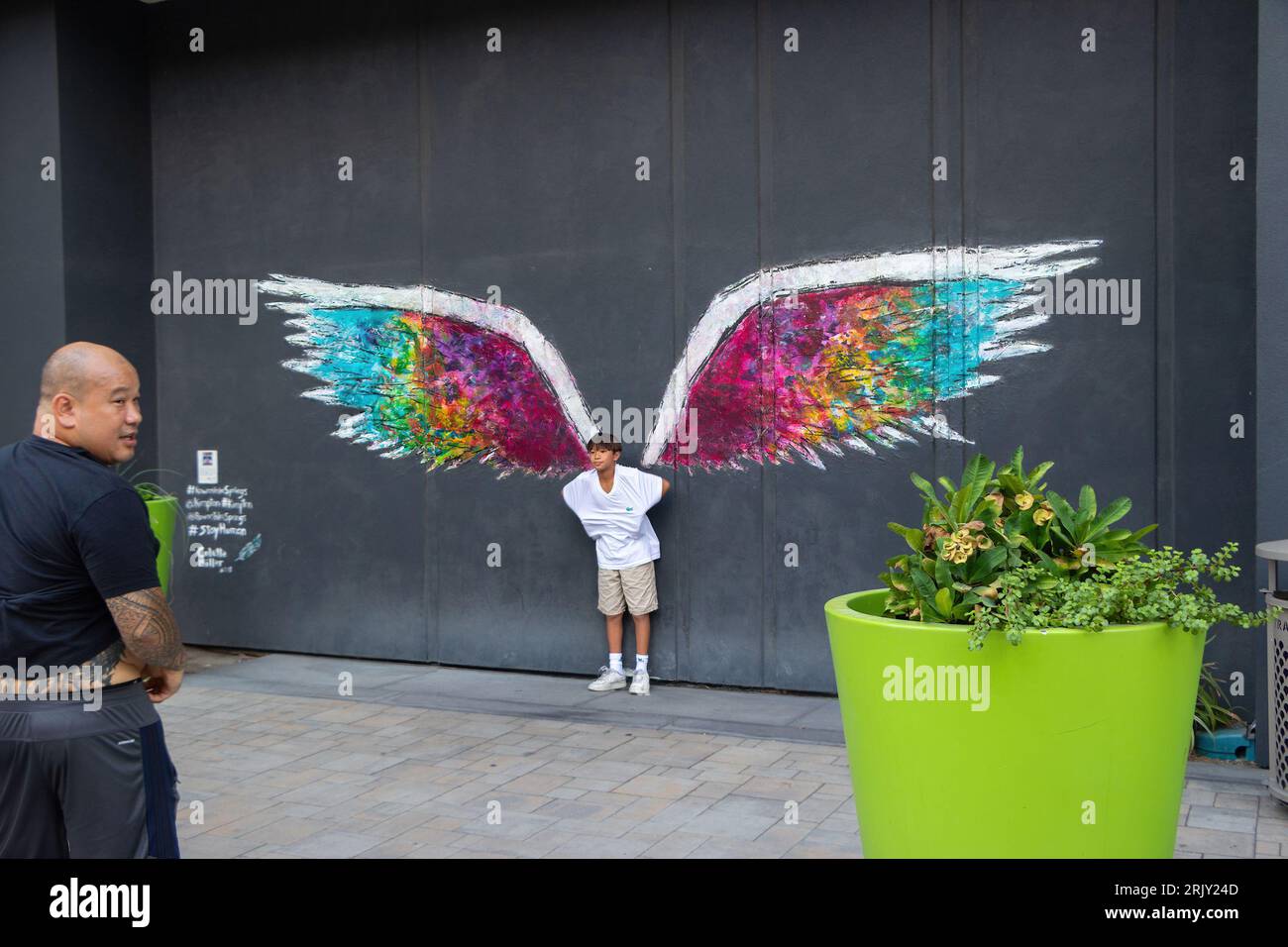 Un ragazzo asiatico si trova di fronte al colorato murale dell'ala dell'angelo su un muro scuro, mentre un uomo guarda sopra, una pianta in vaso verde è nelle vicinanze. palm springs, california Foto Stock