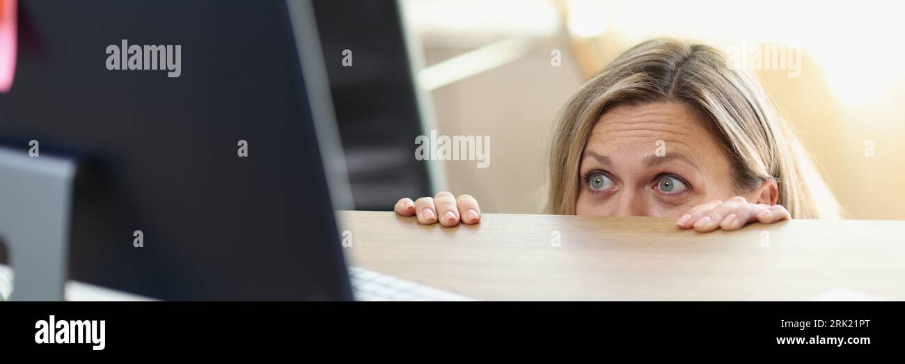 Spaventato manager femminile nascosto dietro la scrivania dell'ufficio e guardando lo schermo del computer. Foto Stock