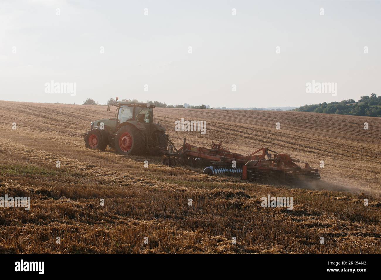 Un trattore moderno con un pesante erpice a disco trainato ara un campo di grano al tramonto. Foto Stock