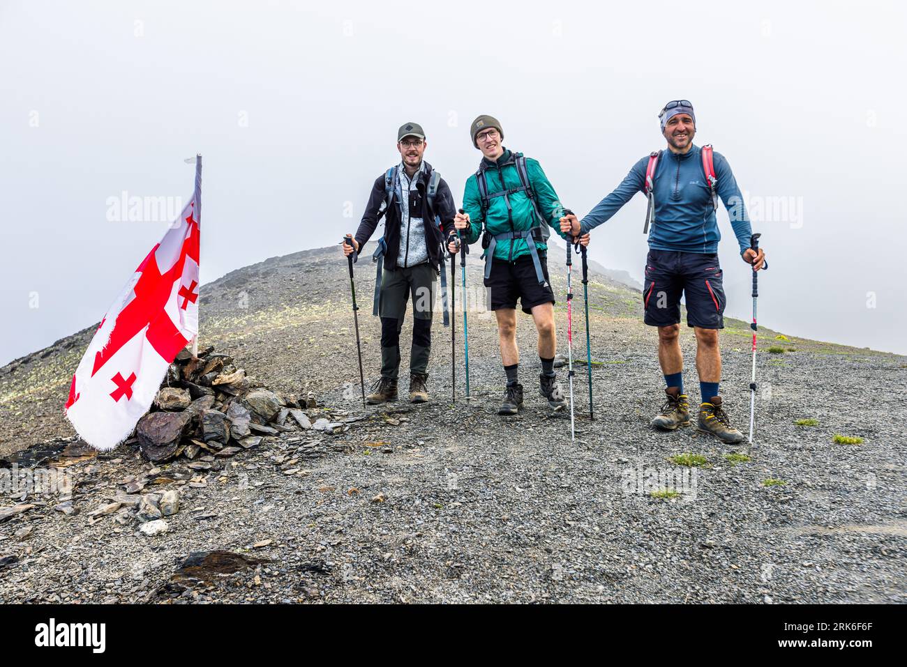 Escursione sul Passo Chaukhi nella Municipalità di Dusheti, regione Mtskheta-Mtianeti, Georgia. Moritz (a sinistra) e Julius (al centro), figli del fotografo Georg Berg, stanno con la guida escursionistica Giorgi Chachua (a destra) in cima al passo accanto a una bandiera georgiana. La bandiera bianca con la croce rossa di San Giorgio e quattro piccole croci rosse è conficcata in un mucchio di pietre. Tutti e tre si appoggiano su bastoni da trekking. Moritz indossa un cappello, occhiali e un gilet grigio sopra una giacca scura. Julius indossa una giacca verde e un cappello di lana. Giorgi Chachua indossa un foulard e una camicia azzurra a maniche lunghe. La nebbia avvolge lo sfondo. Il p Foto Stock