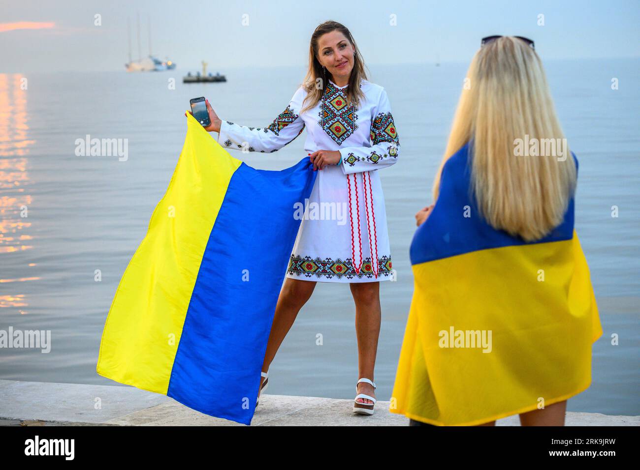 Trieste, Italia, 24 agosto 2023. Le donne ucraine esibiscono la loro bandiera nazionale per celebrare il 32° giorno dell'indipendenza dell'Ucraina nel porto Adriatico di Trieste, Italia. I residenti ucraini hanno tenuto una manifestazione nella centrale Piazza Unità d'Italia facevano parte di una "catena di unità" organizzata dalle comunità ucraine di tutto il mondo per mostrare la loro resilienza, dignità e sostegno al loro paese, invasa dalla Russia esattamente 18 mesi fa. Lontano all'orizzonte si trova il megayacht più grande del mondo, lo yacht a vela da 530 milioni di euro appartenente all'oligarca russo Andrey Igorevich Melnichenko, sequestrato il 20 marzo Foto Stock