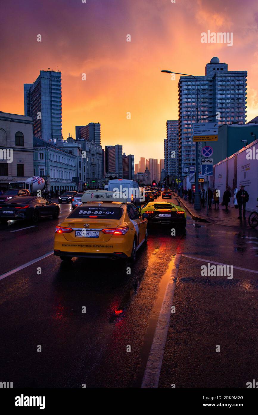 La via Arbat di Mosca prende vita sotto l'ipnotico cielo del tramonto, i suoi edifici storici sovietici e il traffico intenso che si crogiolano nel bagliore post-pioggia. Foto Stock