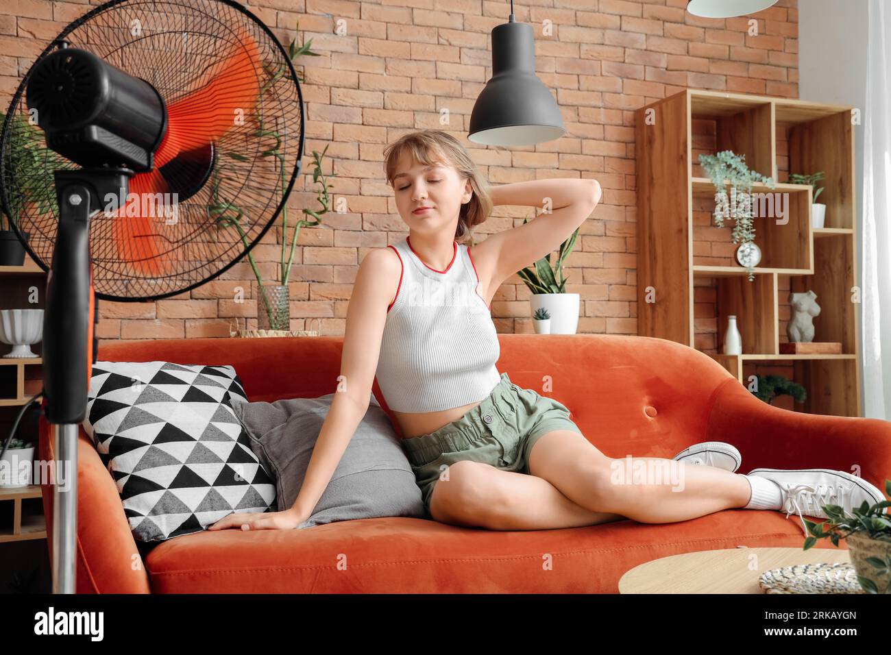 Giovane donna con ventilatore elettrico che soffia seduto sul divano a casa  Foto stock - Alamy