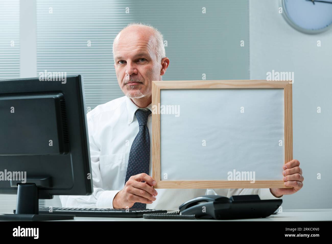 uomo d'affari anziano con maglietta bianca e cravatta siede a una scrivania con monitor, tastiera e telefono. Tenendo un segno bianco, il suo sguardo serio suggerisce professi Foto Stock